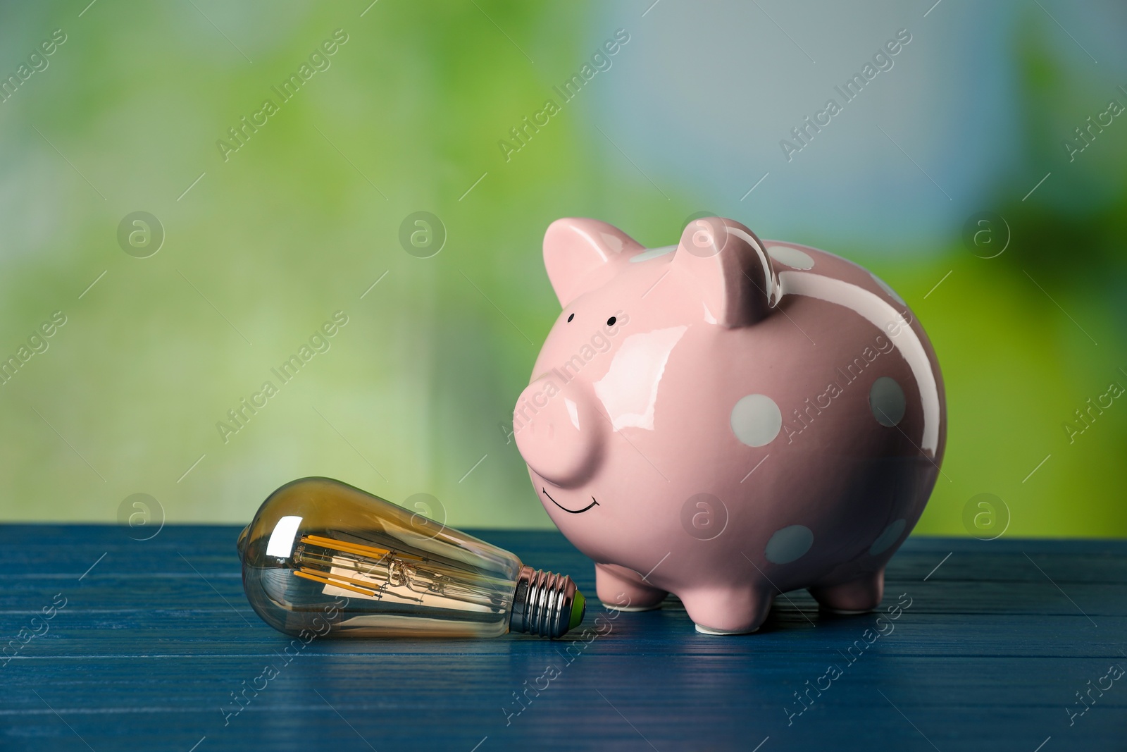 Photo of Piggy bank and light bulb on blue wooden table outdoors. Energy saving concept