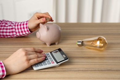 Photo of Woman putting coin into piggy bank while using calculator at wooden table, closeup. Energy saving concept