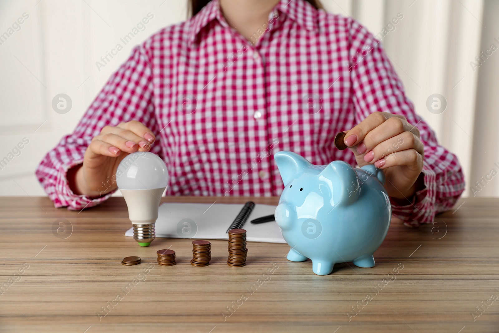 Photo of Woman with light bulb putting coin into piggy bank at wooden table, closeup. Energy saving concept