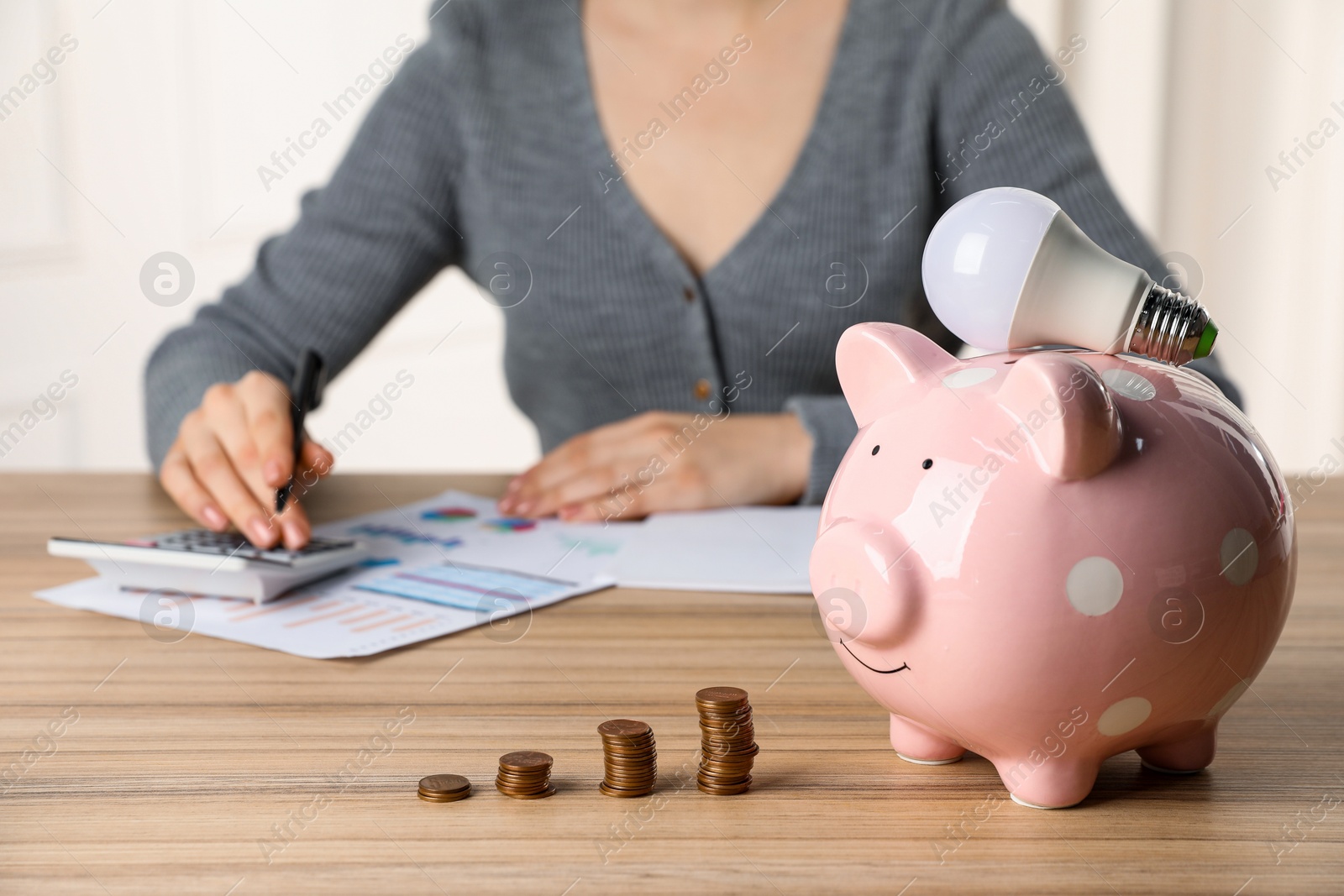 Photo of Woman using calculator at wooden table, focus on piggy bank, coins and light bulb. Energy saving concept