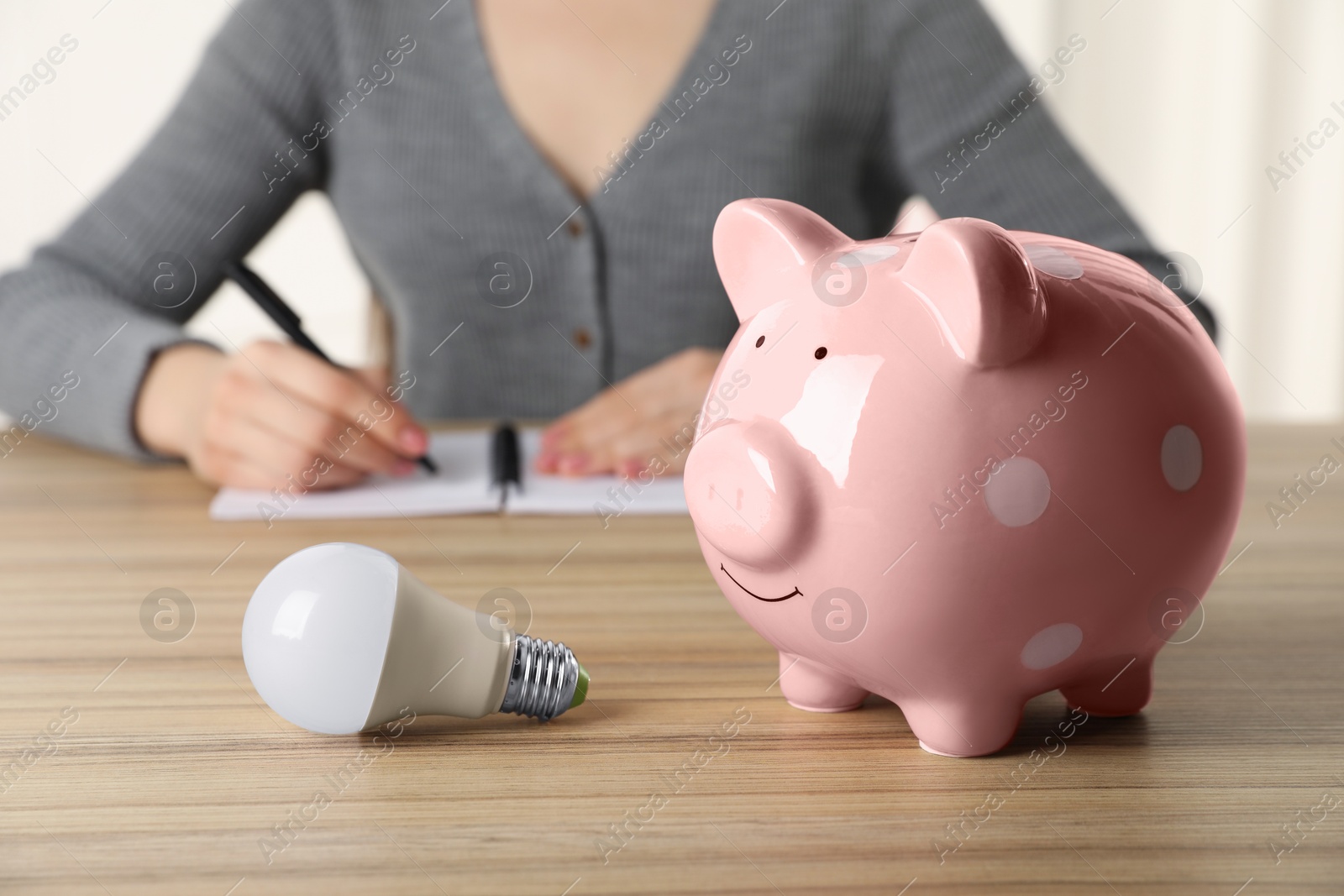 Photo of Woman taking notes at wooden table, focus on piggy bank and light bulb. Energy saving concept