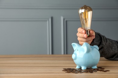 Photo of Energy saving concept. Man holding light bulb above piggy bank at wooden table, closeup. Space for text