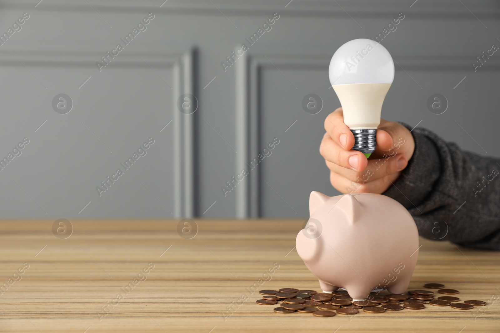 Photo of Energy saving concept. Man holding light bulb above piggy bank at wooden table, closeup. Space for text