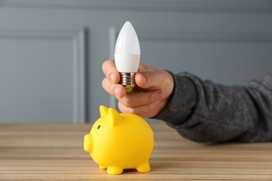 Photo of Man holding light bulb above piggy bank at wooden table, closeup. Energy saving concept