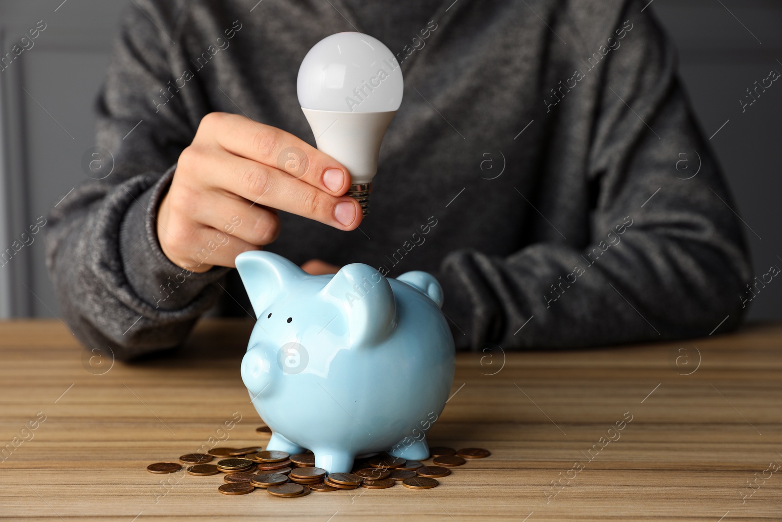 Photo of Man holding light bulb above piggy bank at wooden table, closeup. Energy saving concept