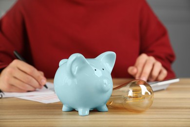 Photo of Man taking notes while using calculator at wooden table, focus on piggy bank and light bulb. Energy saving concept