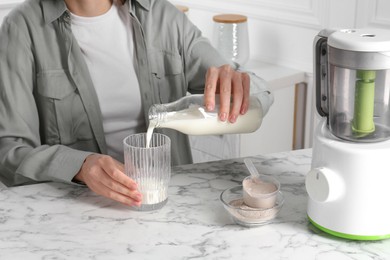 Photo of Making protein cocktail. Woman pouring milk from bottle into glass at white marble table, closeup