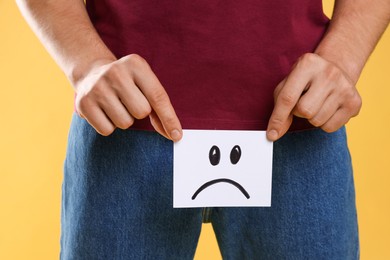 Photo of STD (sexually transmitted diseases). Man holding sticky note with drawn sad face on yellow background, closeup