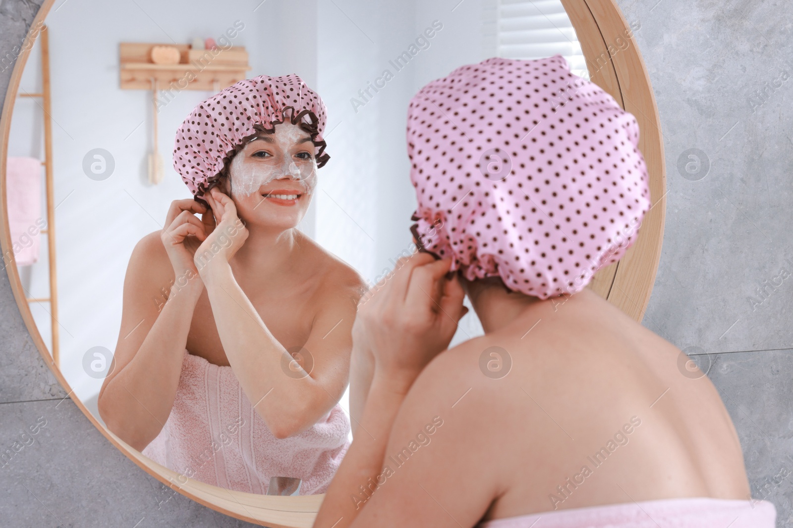 Photo of Woman with shower cap and cream on her face near mirror in bathroom