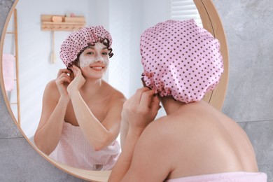 Photo of Woman with shower cap and cream on her face near mirror in bathroom