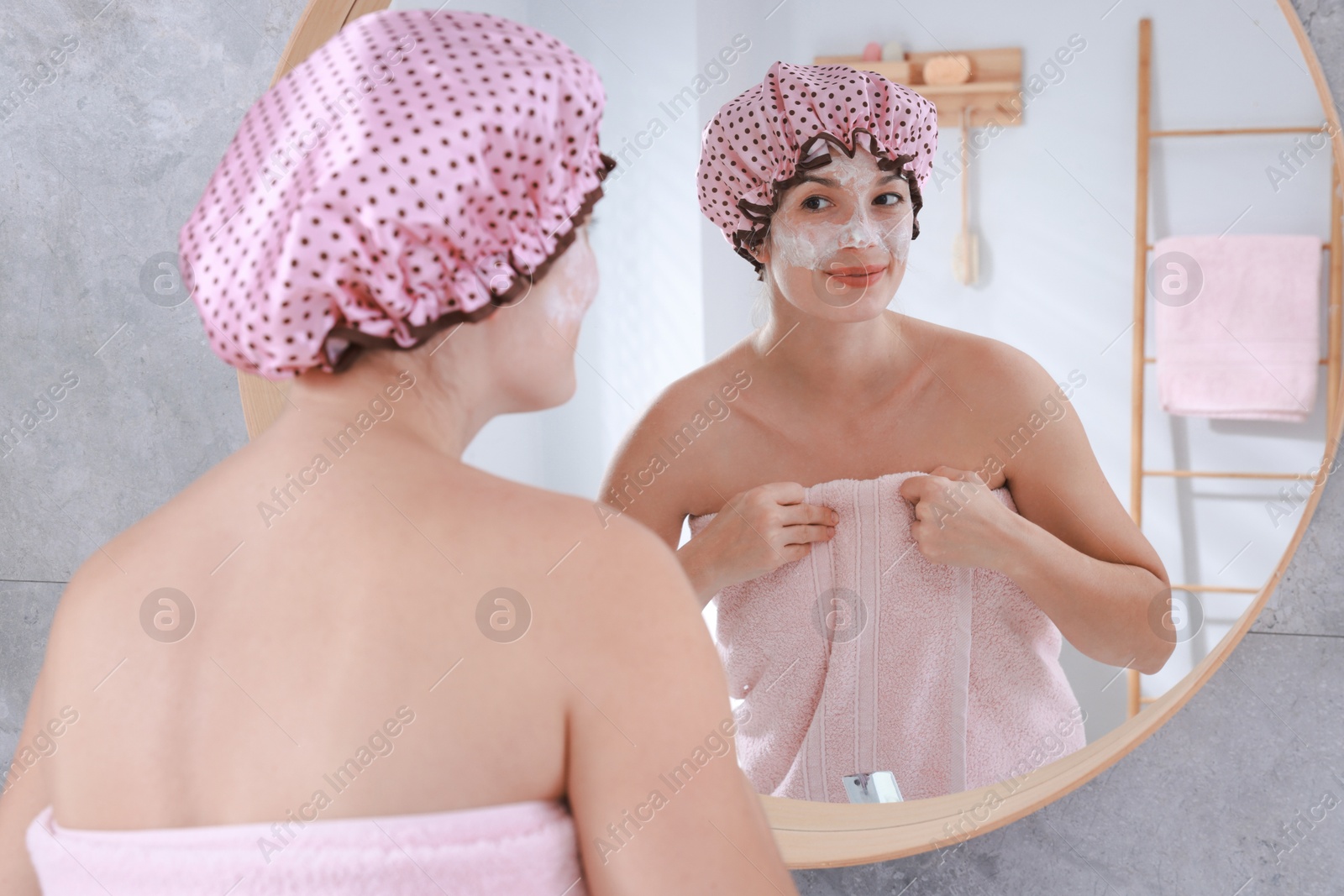 Photo of Woman with shower cap and cream on her face near mirror in bathroom