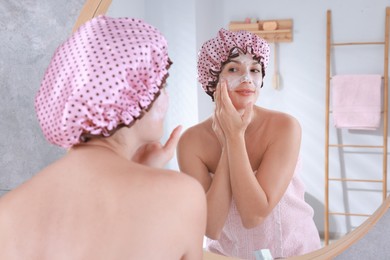 Photo of Woman with shower cap applying face cream near mirror in bathroom