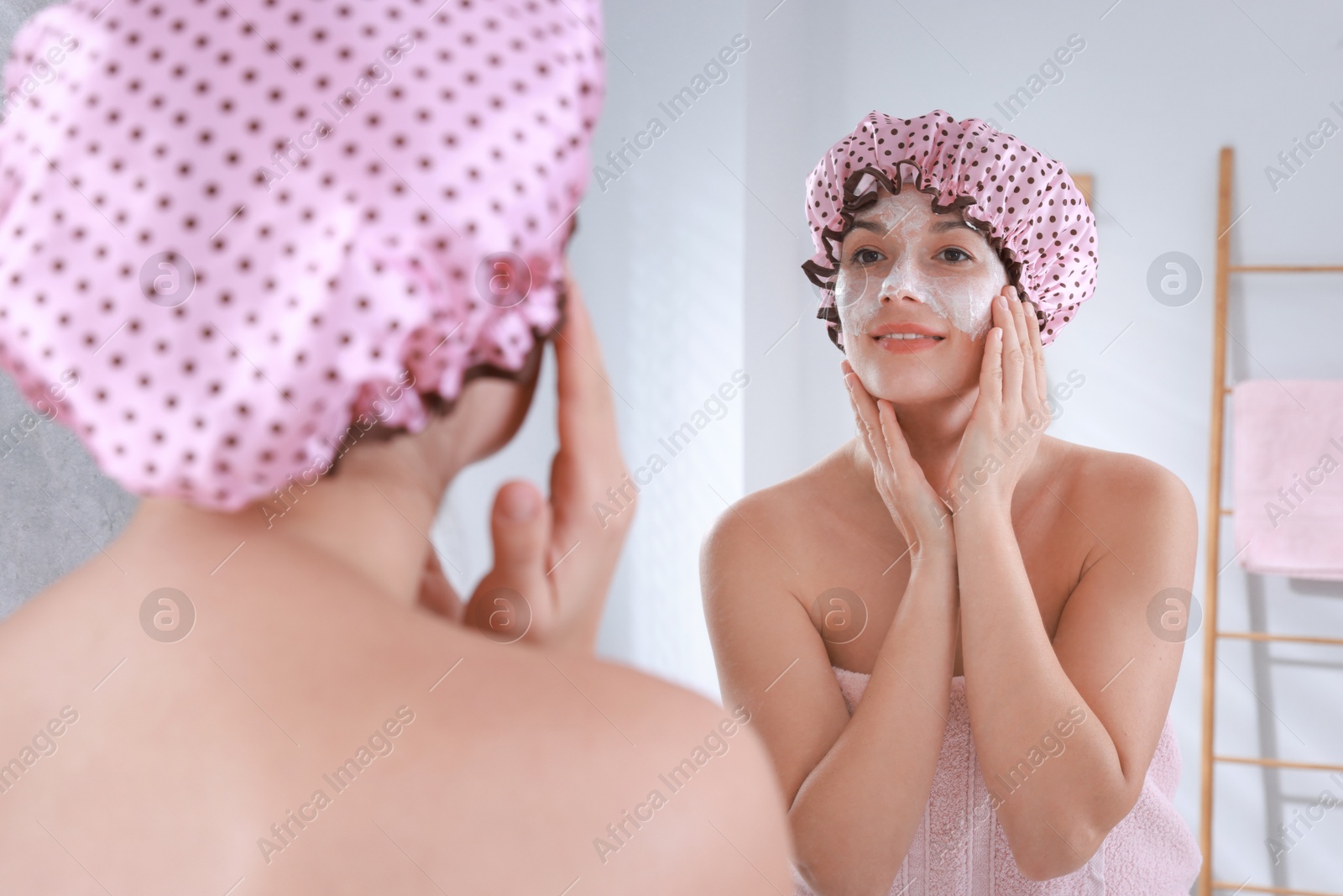 Photo of Woman with shower cap applying face cream near mirror in bathroom