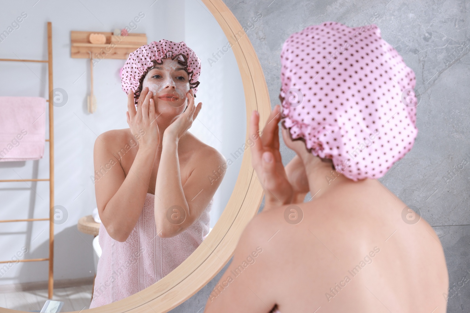 Photo of Woman with shower cap applying face cream near mirror in bathroom