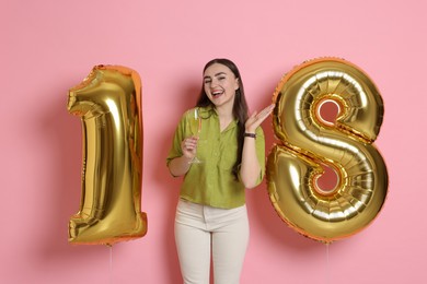 Photo of Coming of age party - 18th birthday. Happy young woman with number shaped balloons and glass of sparkling wine on pink background