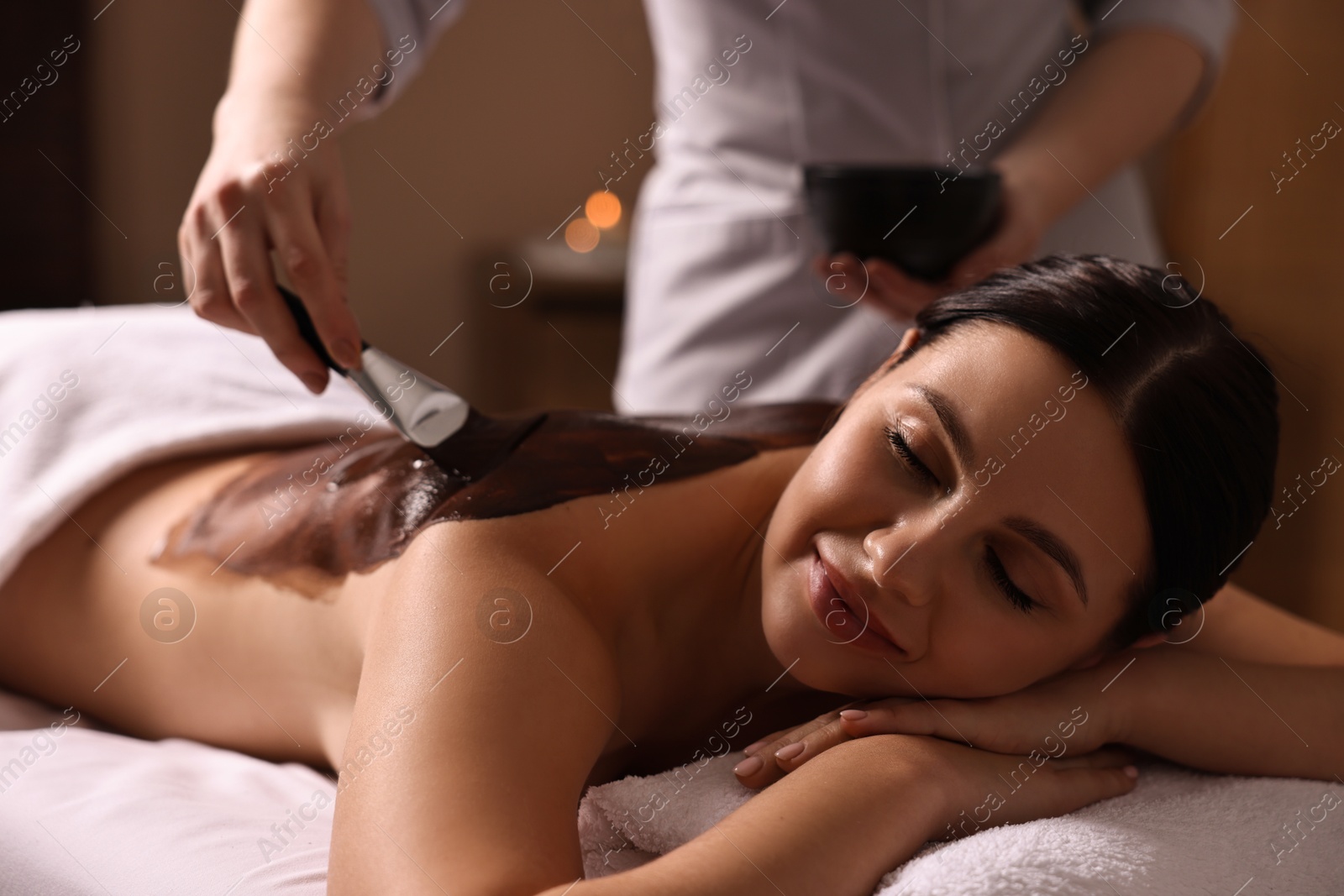 Photo of Chocolate body wrap. Spa worker applying mask onto woman's back in salon, closeup