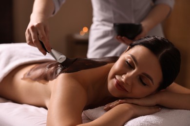 Photo of Chocolate body wrap. Spa worker applying mask onto woman's back in salon, closeup