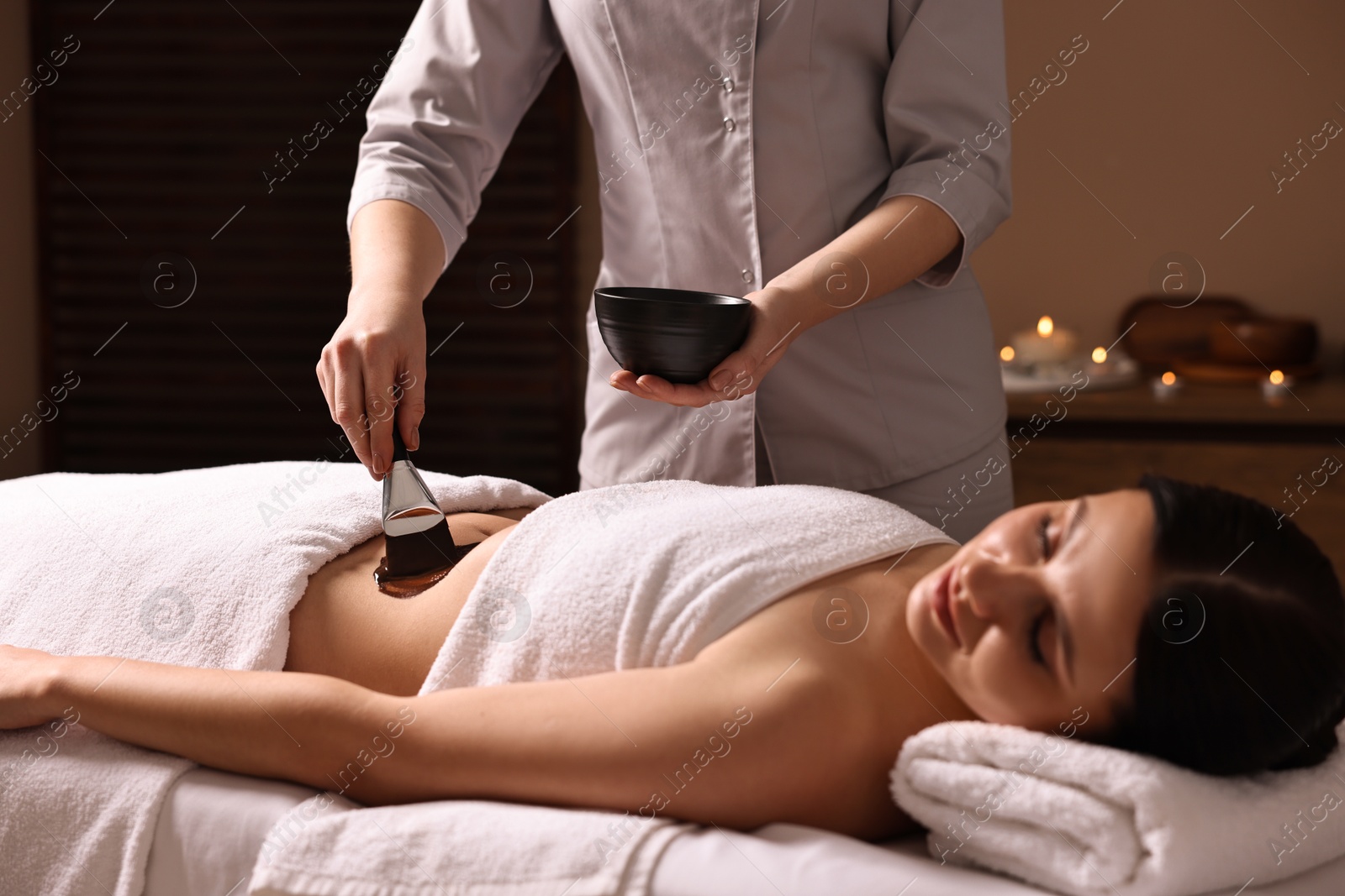 Photo of Chocolate body wrap. Spa worker applying mask onto woman's belly in salon, closeup