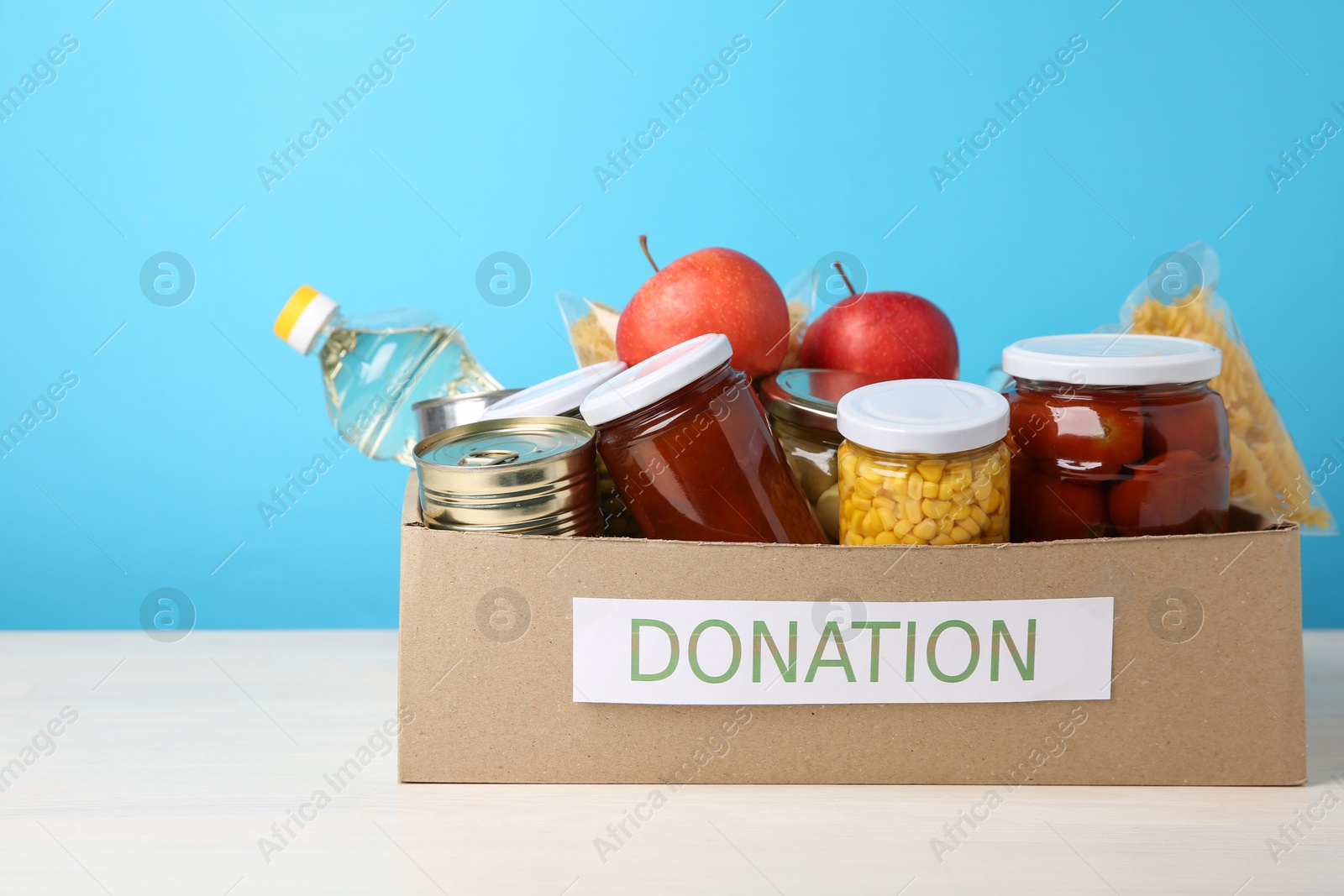 Photo of Different food products for donation in box on table against light blue background