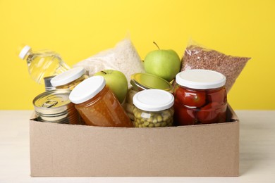 Photo of Different food products for donation in box on table against yellow background