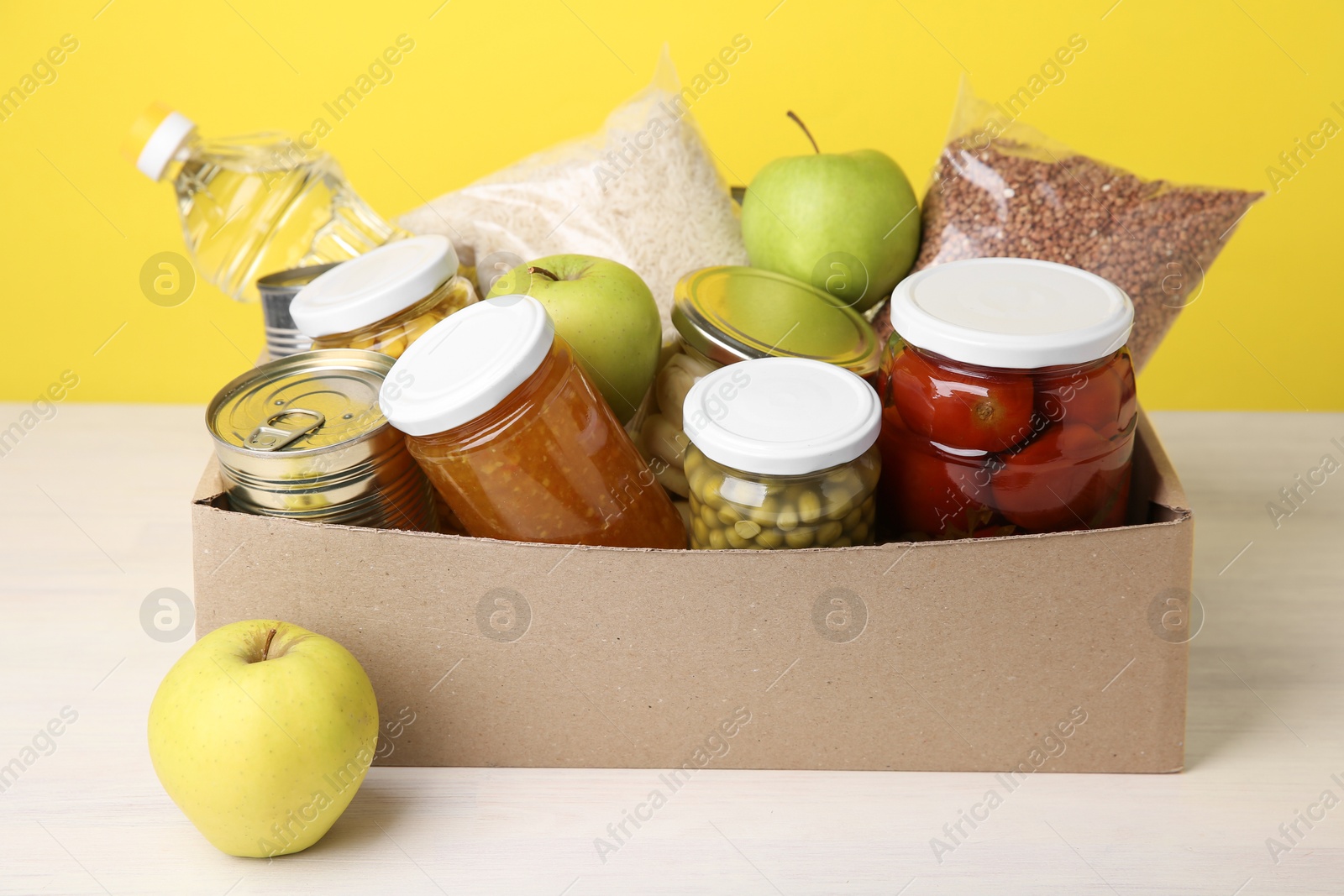 Photo of Different food products for donation in box on table against yellow background