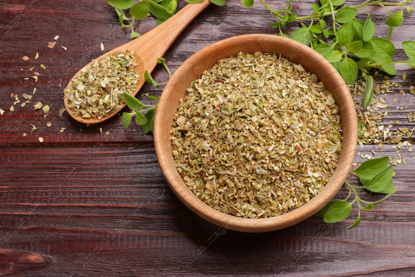 Photo of Dried oregano in bowl, spoon and green leaves on wooden table, top view