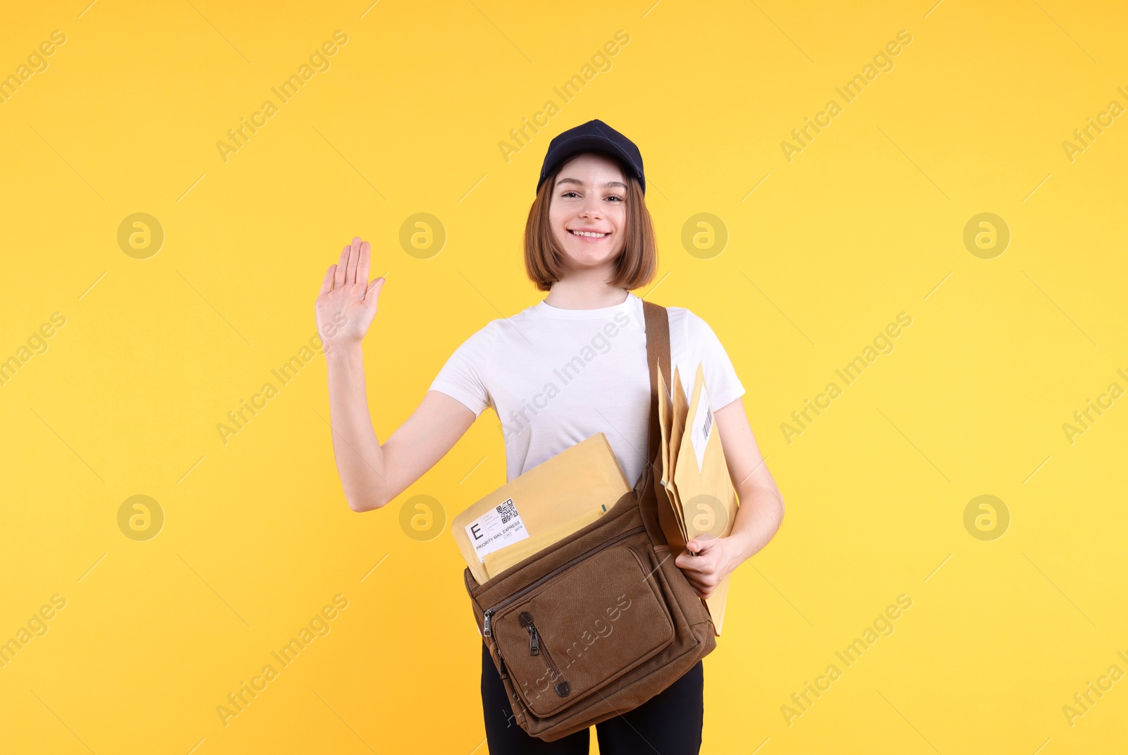 Photo of Happy postwoman with bag and envelopes waving hello on yellow background. Space for text