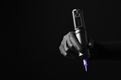 Photo of Professional artist in gloves holding tattoo machine on black background, closeup. Space for text