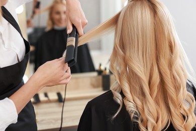 Photo of Hairdresser curling woman's hair with flat iron in salon, closeup