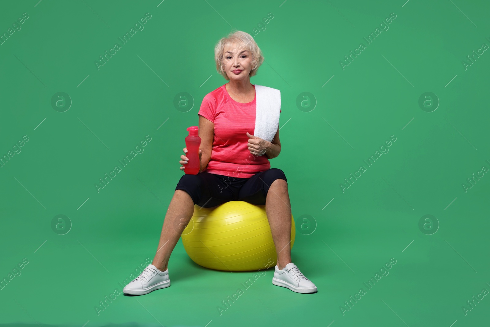 Photo of Senior woman with fitness ball, water and towel on green background