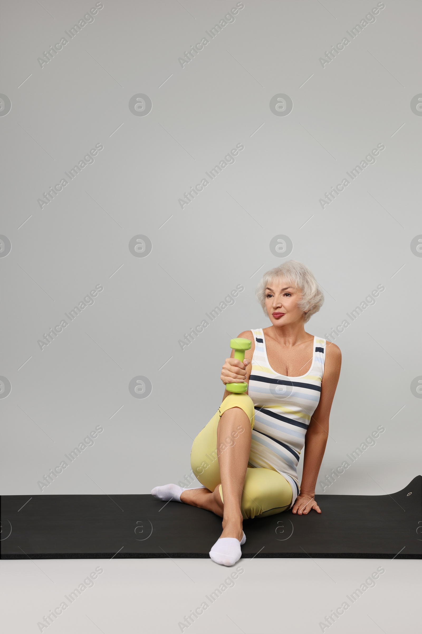 Photo of Senior woman exercising with fitness mat and dumbbell on light grey background, space for text