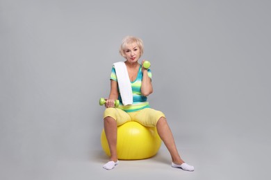 Photo of Senior woman exercising with fitness ball, dumbbells and towel on light grey background