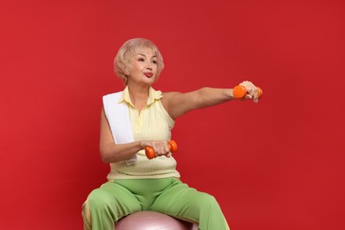 Photo of Senior woman exercising with fitness ball, towel and dumbbells on red background