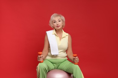 Photo of Senior woman with fitness ball, towel and dumbbells on red background