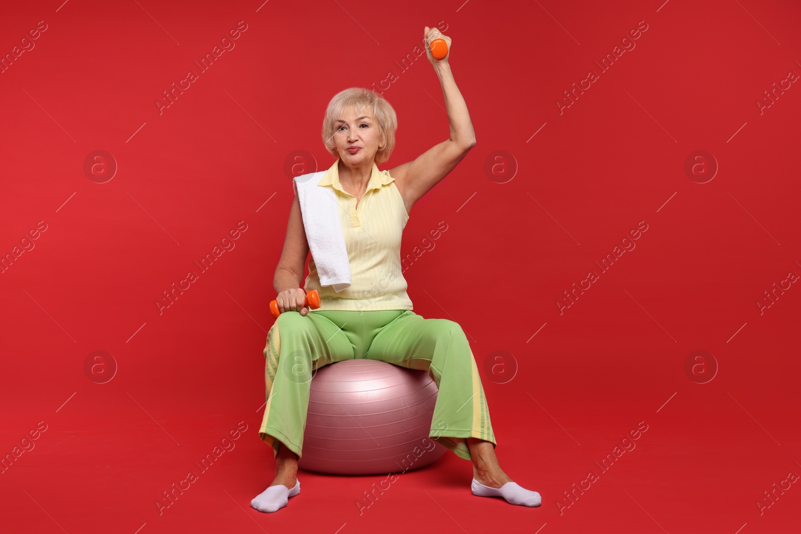 Photo of Senior woman exercising with fitness ball, towel and dumbbells on red background