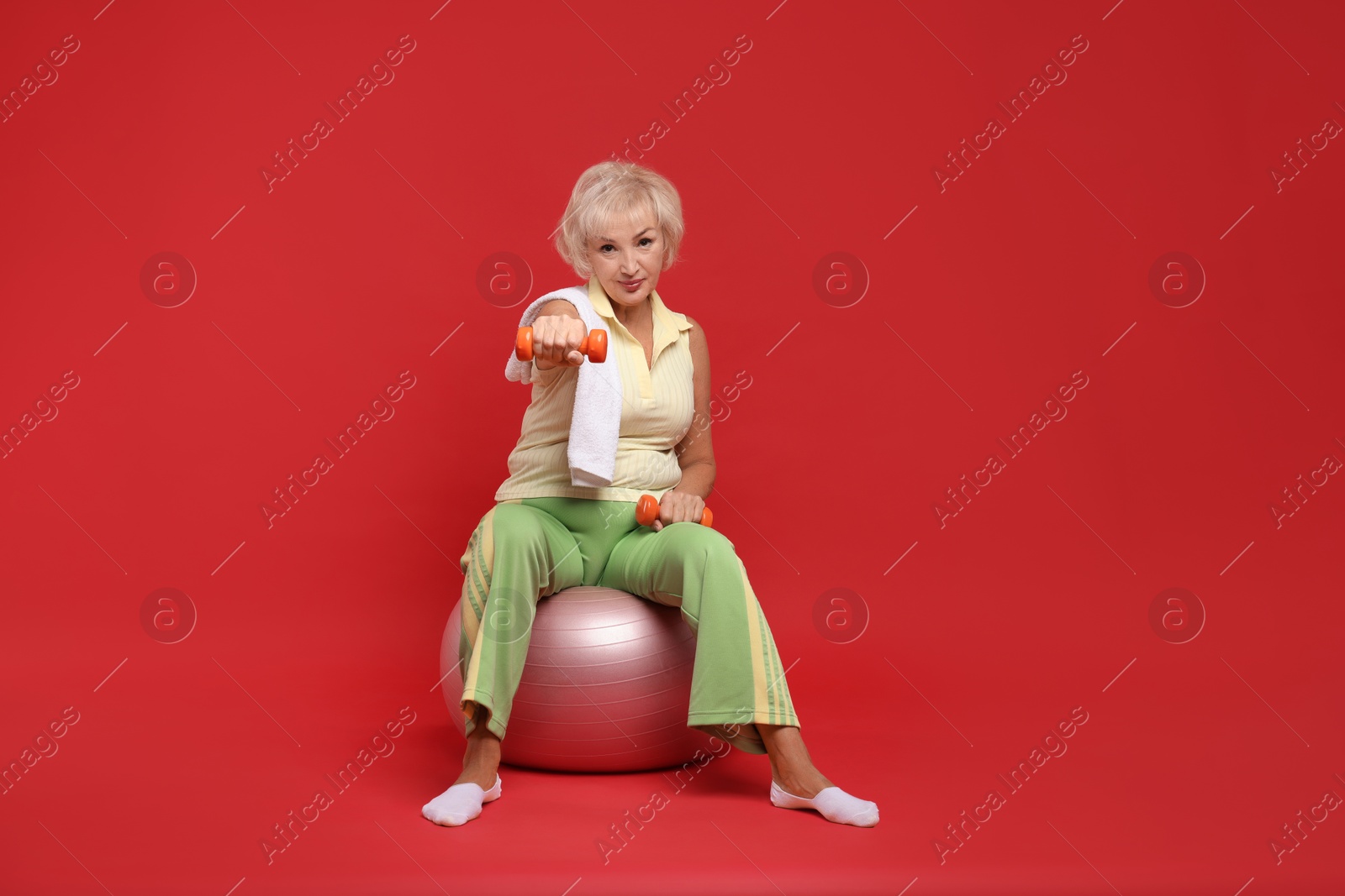 Photo of Senior woman exercising with fitness ball, towel and dumbbells on red background