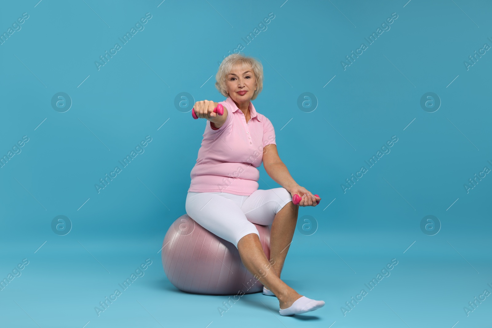 Photo of Senior woman exercising with fitness ball and dumbbells on light blue background