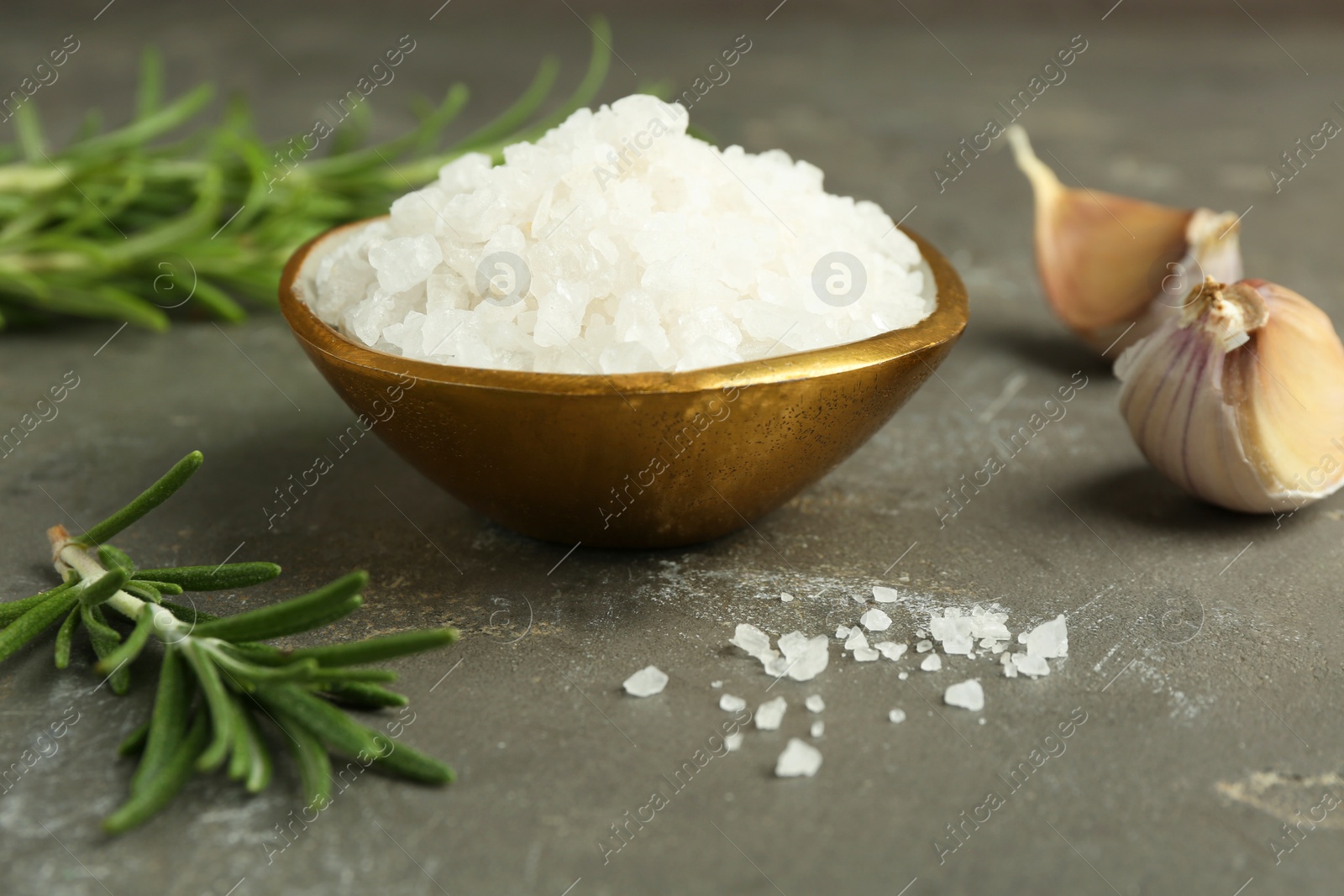 Photo of Sea salt in bowl, rosemary and garlic on grey table, closeup