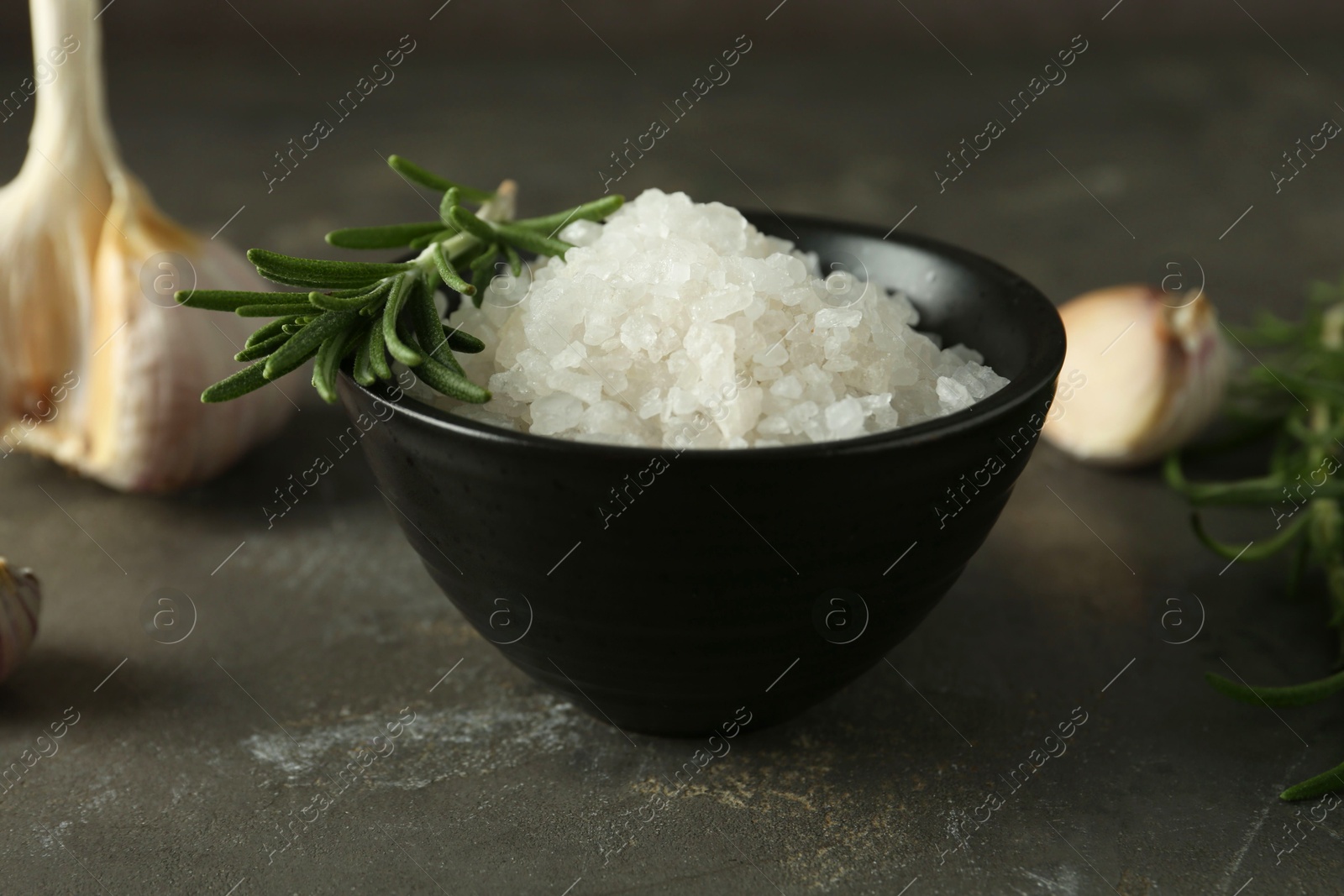 Photo of Sea salt in bowl, rosemary and garlic on grey table, closeup