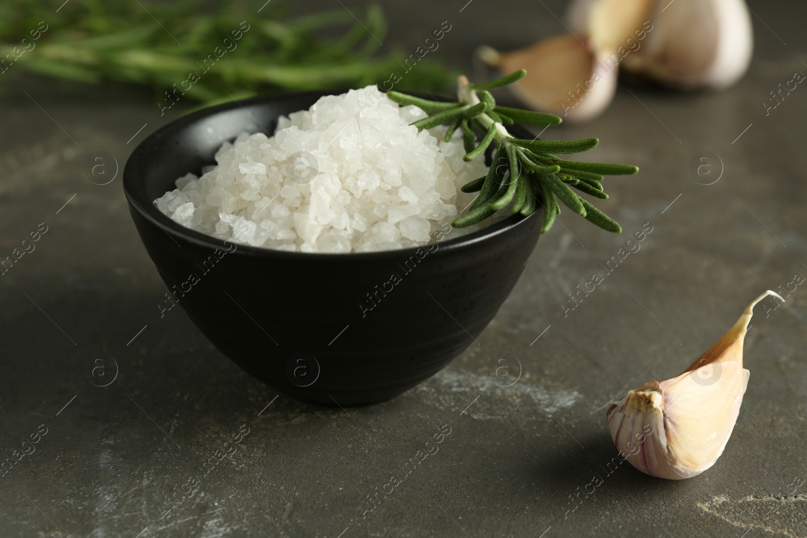 Photo of Sea salt in bowl, rosemary and garlic on grey table, closeup