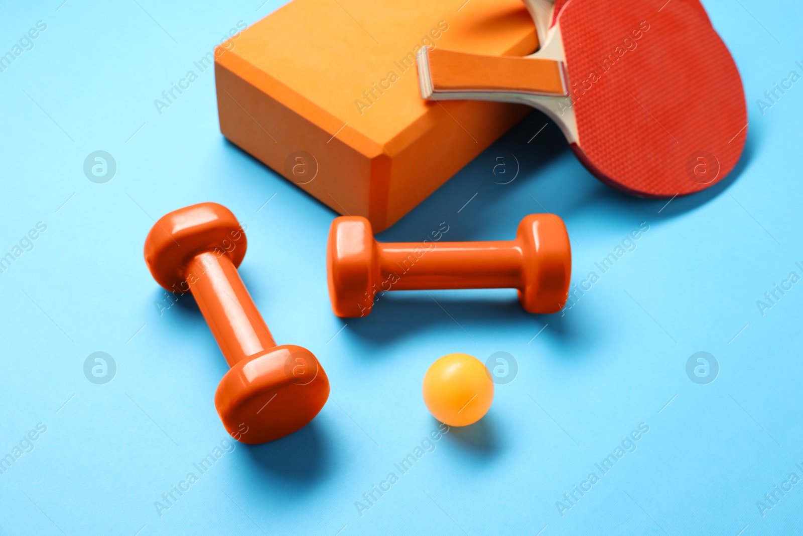 Photo of Dumbbells, yoga block, ping pong rackets and ball on light blue background, closeup