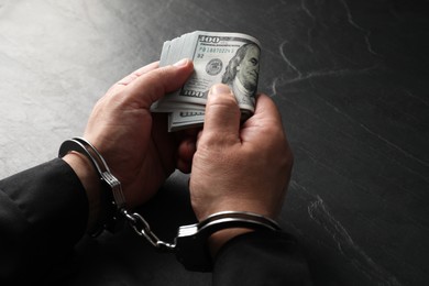 Photo of Corruption. Man in handcuffs holding dollar banknotes at grey textured table, closeup