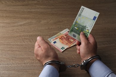 Photo of Corruption. Man in handcuffs holding euro banknotes at wooden table, closeup