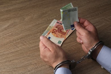 Photo of Corruption. Man in handcuffs holding euro banknotes at wooden table, closeup