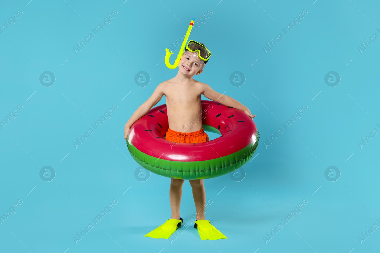 Photo of Cute little boy in beachwear with snorkeling equipment and inflatable ring on light blue background