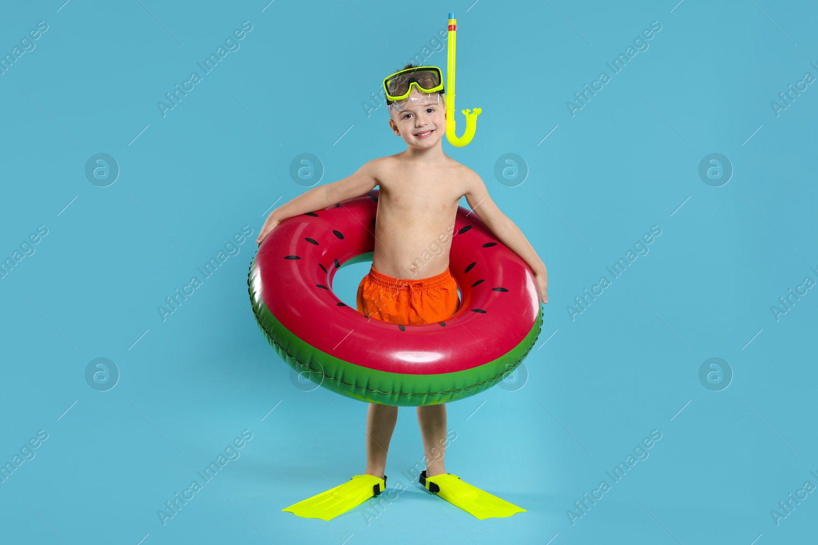 Photo of Cute little boy in beachwear with snorkeling equipment and inflatable ring on light blue background