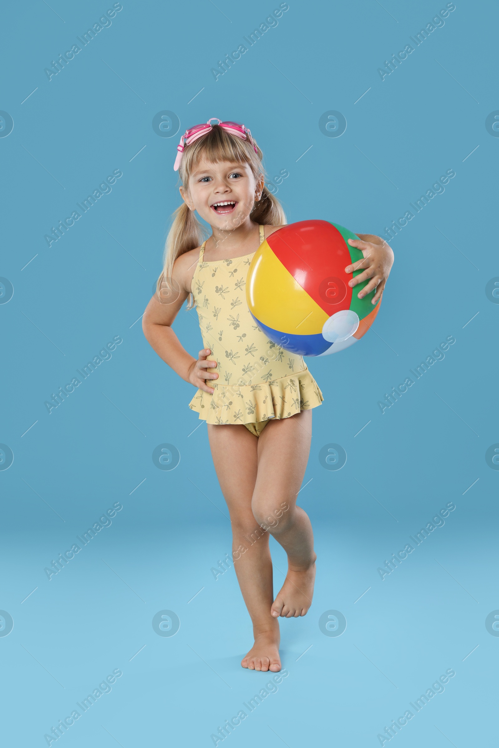 Photo of Cute little girl in swimsuit with inflatable ball and swim goggles on light blue background