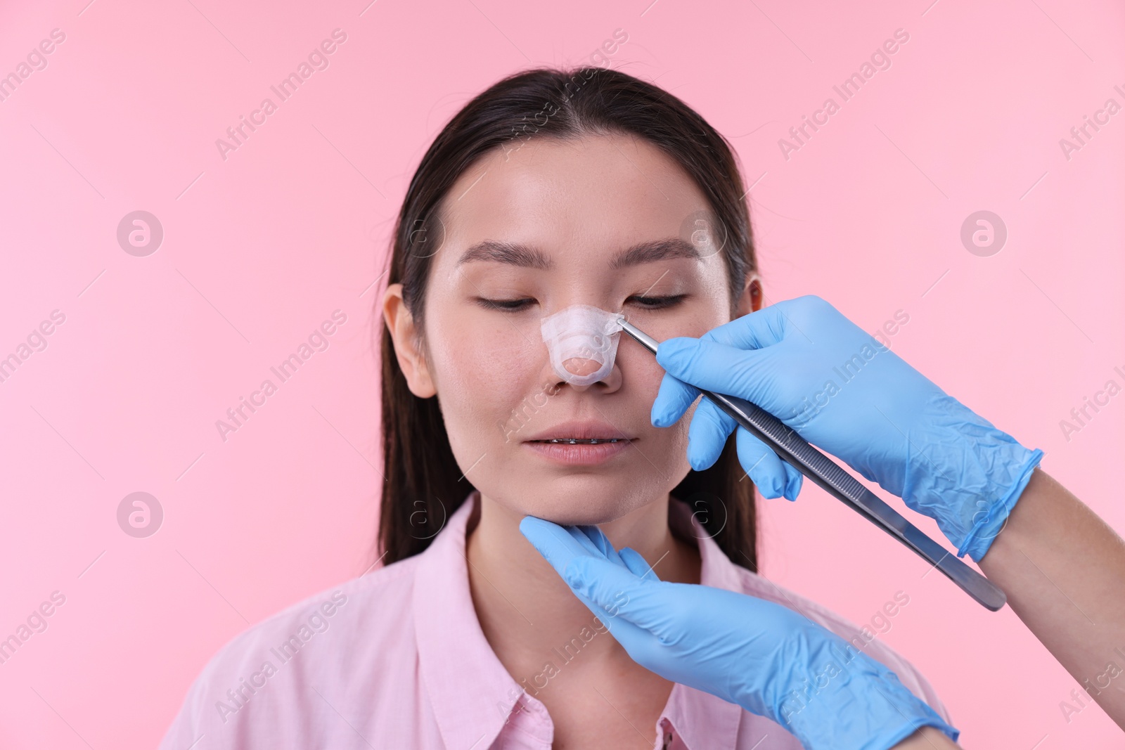 Photo of Doctor removing medical bandage from patient's nose after plastic surgery operation on pink background, closeup