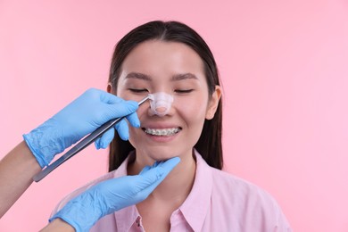 Photo of Doctor removing medical bandage from patient's nose after plastic surgery operation on pink background, closeup