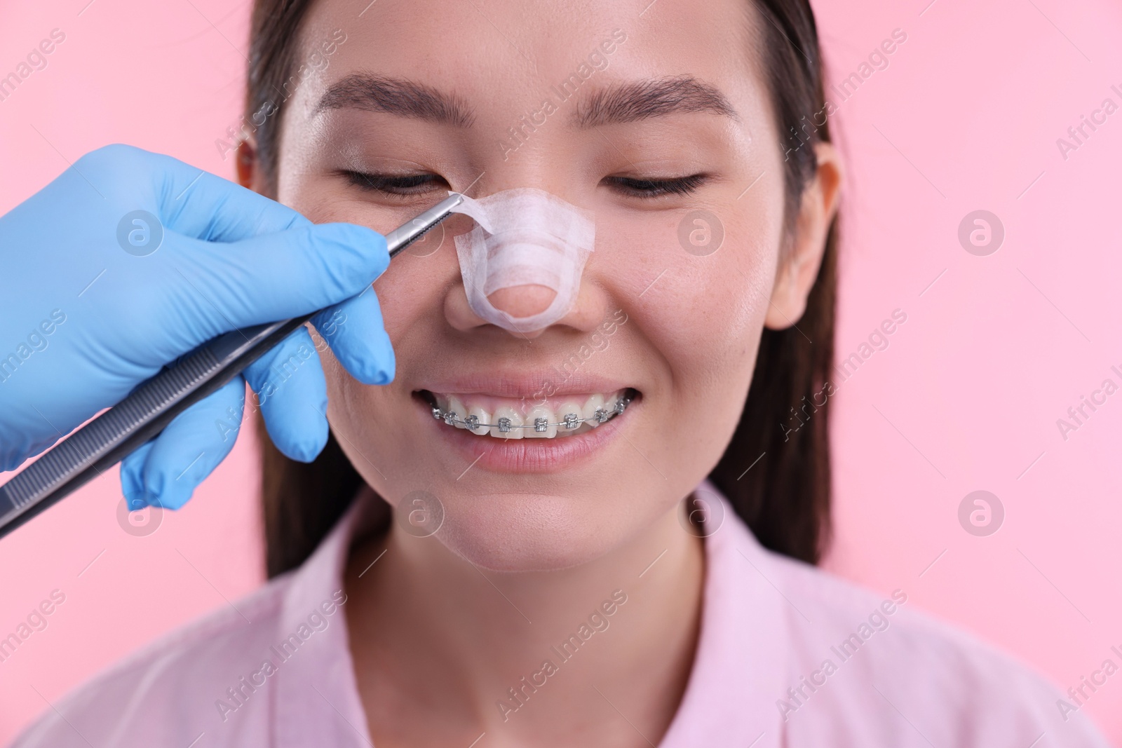 Photo of Doctor removing medical bandage from patient's nose after plastic surgery operation on pink background, closeup